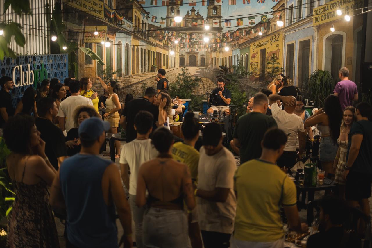 Público lotado dançando no Samba no Quintal do Container 932, com o mural de rua colonial e varal de lâmpadas ao fundo